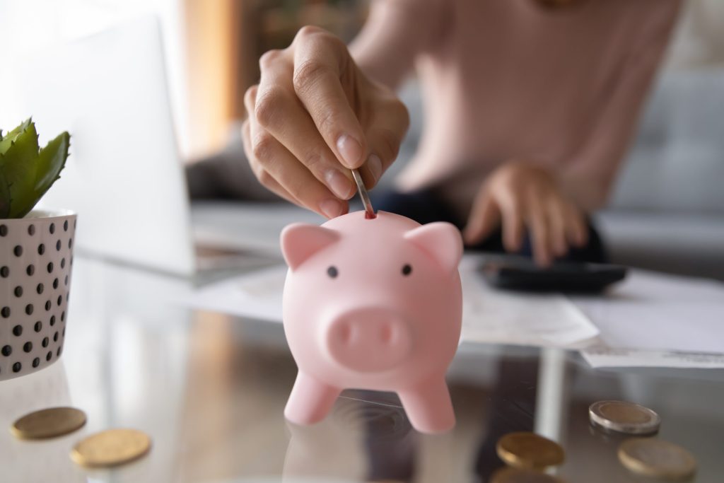 A person’s hand inserts a coin into a pink piggy bank on a glass table with scattered coins and paperwork, symbolizing How to Focus on Your Finances and build better saving habits.