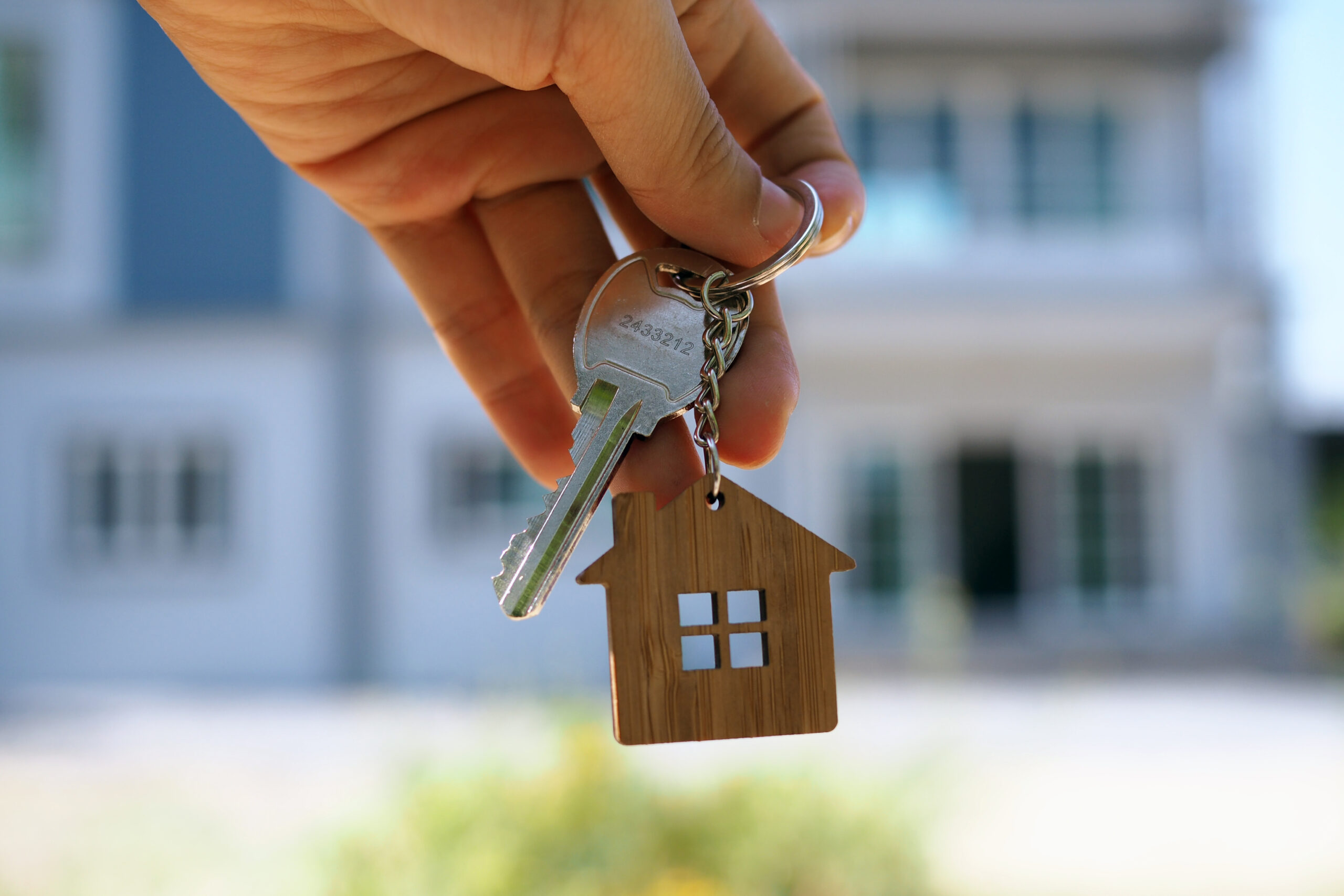 A hand holds a keychain with a silver key and a small wooden house charm, symbolizing the mentorship guidance and decision-making process in selecting a mentor who doesn’t take over, set against a blurred house background.