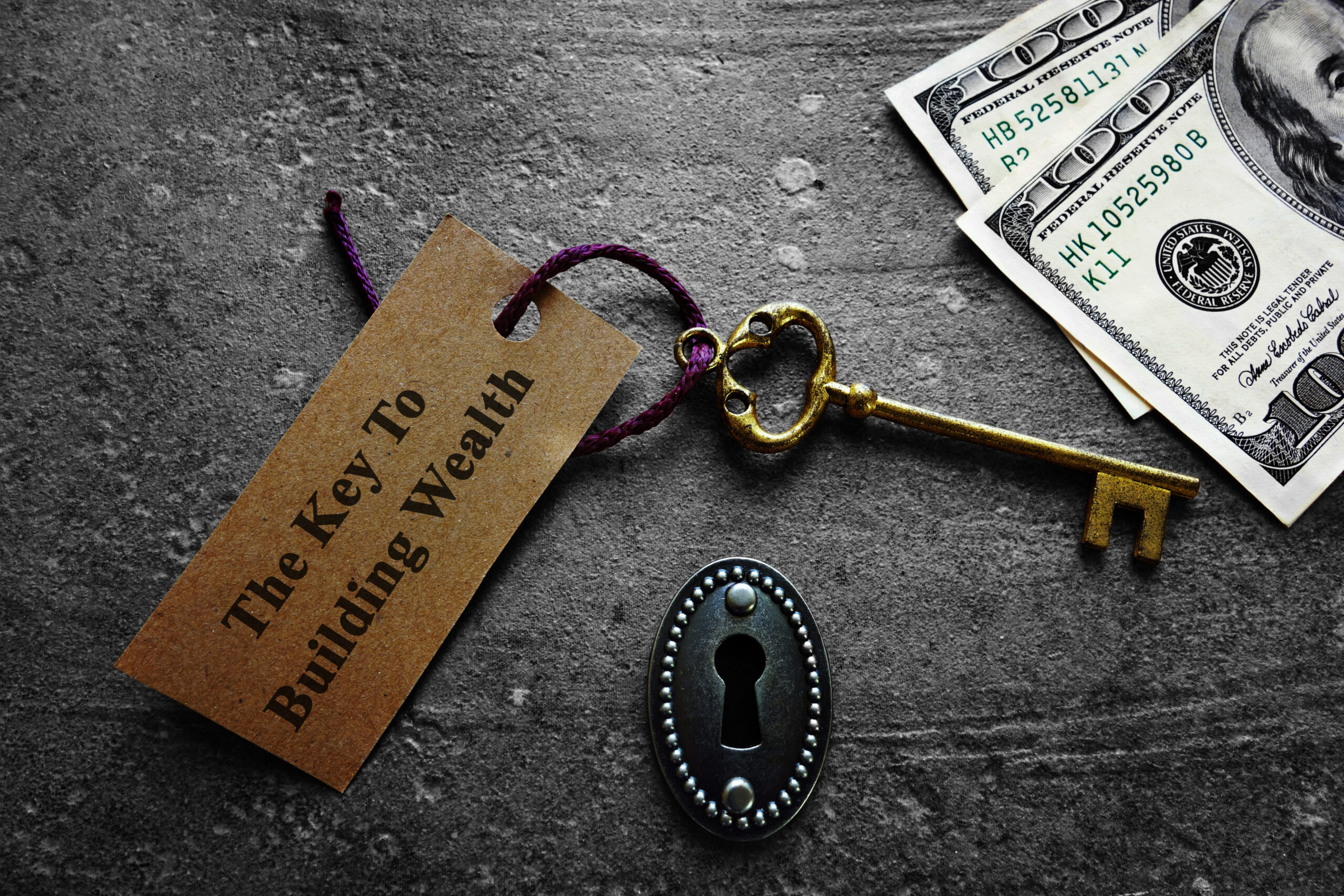 A vintage key with a tag reading The Key To Building Wealth lies next to a keyhole and two hundred-dollar bills, hinting at essential Personal Finance Insights Everyone Needs on a dark, textured surface.