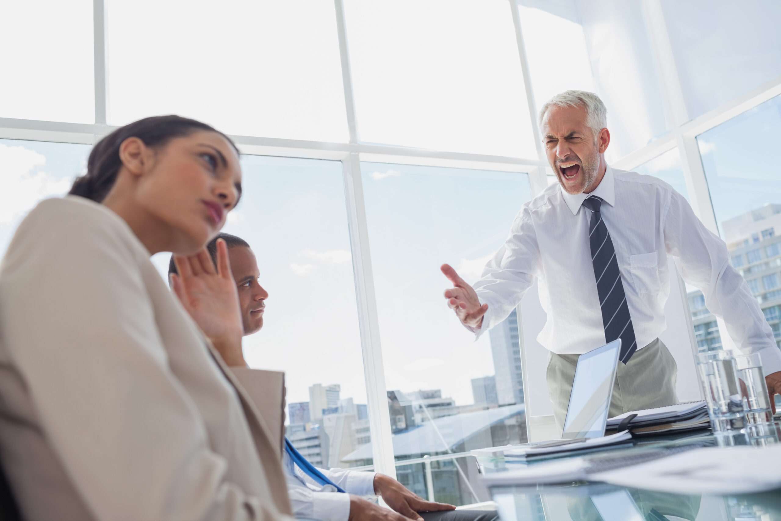 An older man in a white shirt and tie angrily shouts and points at two seated colleagues, showing how bad mentors can create relationship adversity in the office. One woman looks away uncomfortably, while a man beside her appears concerned.