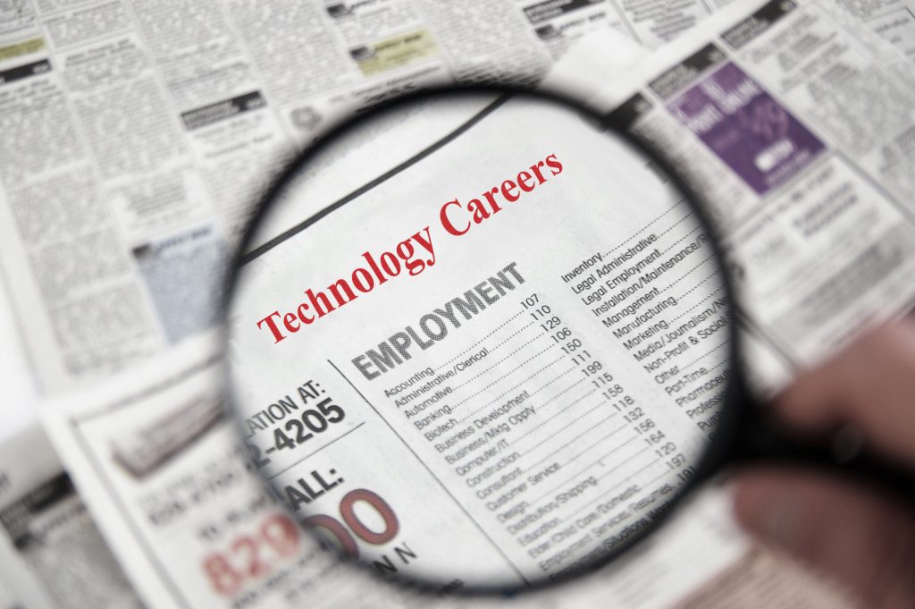 A magnifying glass focuses on a newspaper classified ad with the heading Technology Careers and job listings, while surrounding text blurs—capturing the essence of navigating career transitions with grace in today’s evolving job market.