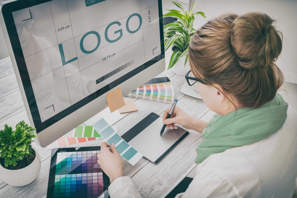 A woman designs a logo on a desktop computer, using a graphics tablet and stylus. She explores color swatches, focusing on personal branding for entrepreneurs to build a powerful connection with her audience. A large monitor displays the word “LOGO.”.