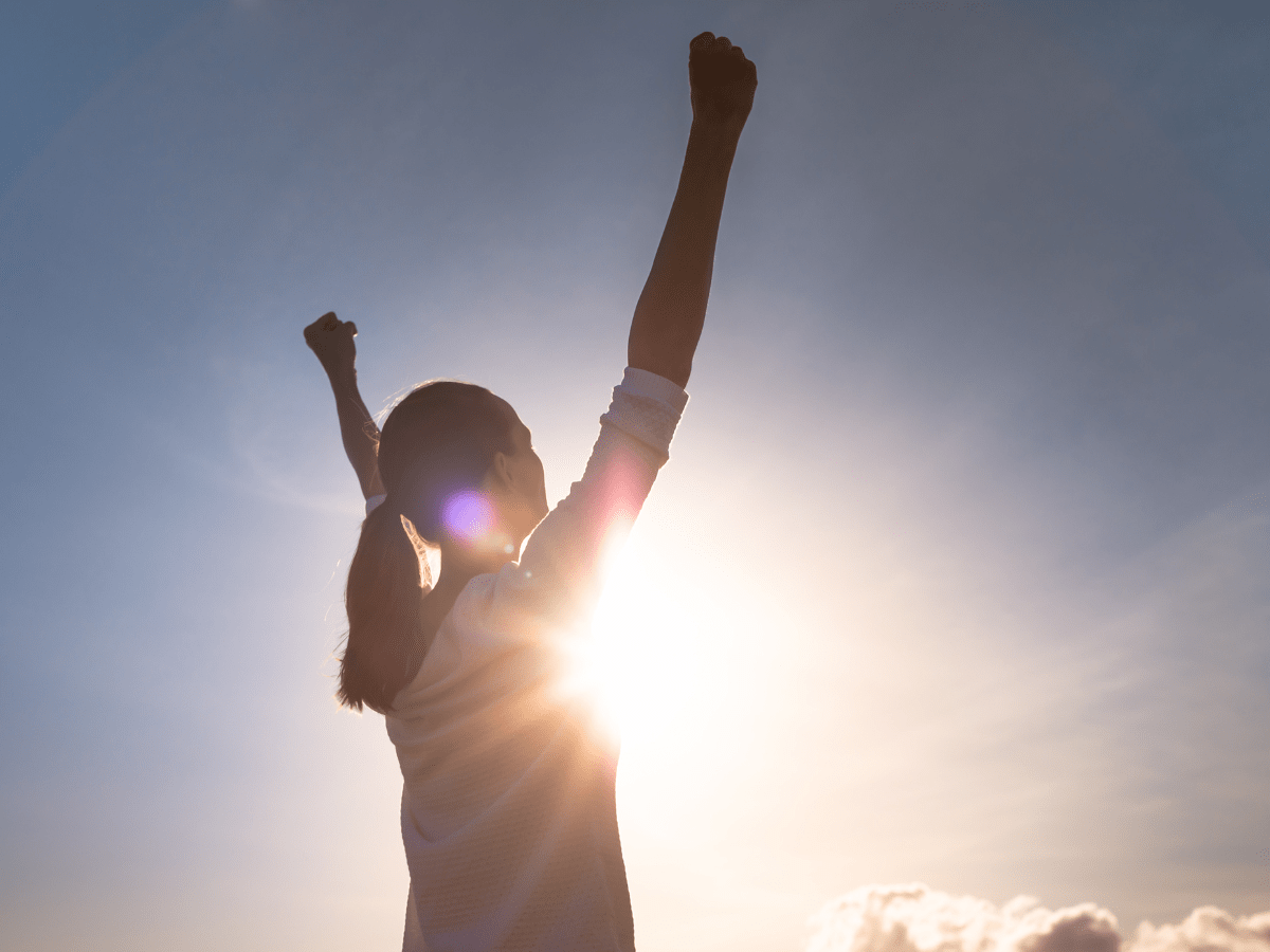A person standing outdoors with arms raised in victory, backlit by the bright sun, embodies how to overcome obstacles and seize opportunities—a dramatic and uplifting silhouette against the sky.