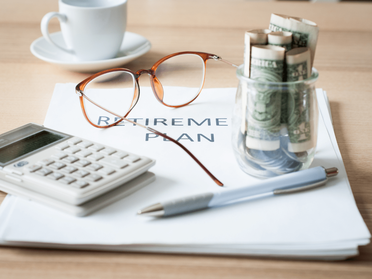 A pair of glasses, a jar of rolled-up cash, a calculator, and a pen rest on a document labeled Retirement Plan—subtle reminders to consider: What is the Best Age to Retire? A coffee cup sits in the background on a light wooden table.