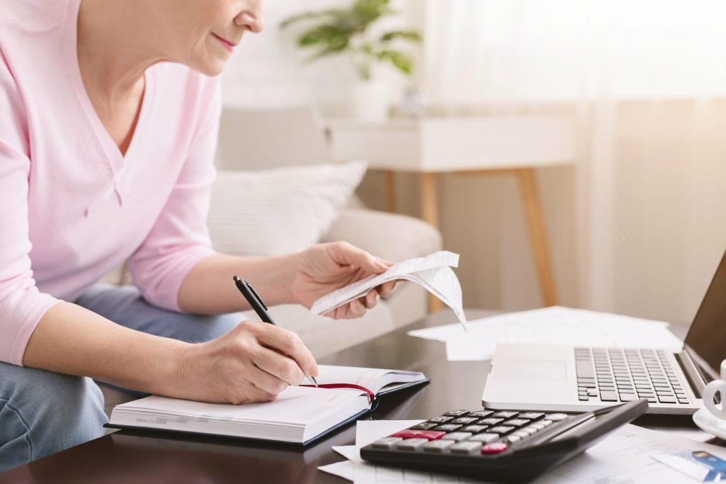 A woman sits at a table reviewing receipts, writing in a notebook, and using a calculator—demonstrating The Most Important Financial Literacy Skills for Women as she makes money work for her while budgeting at home.