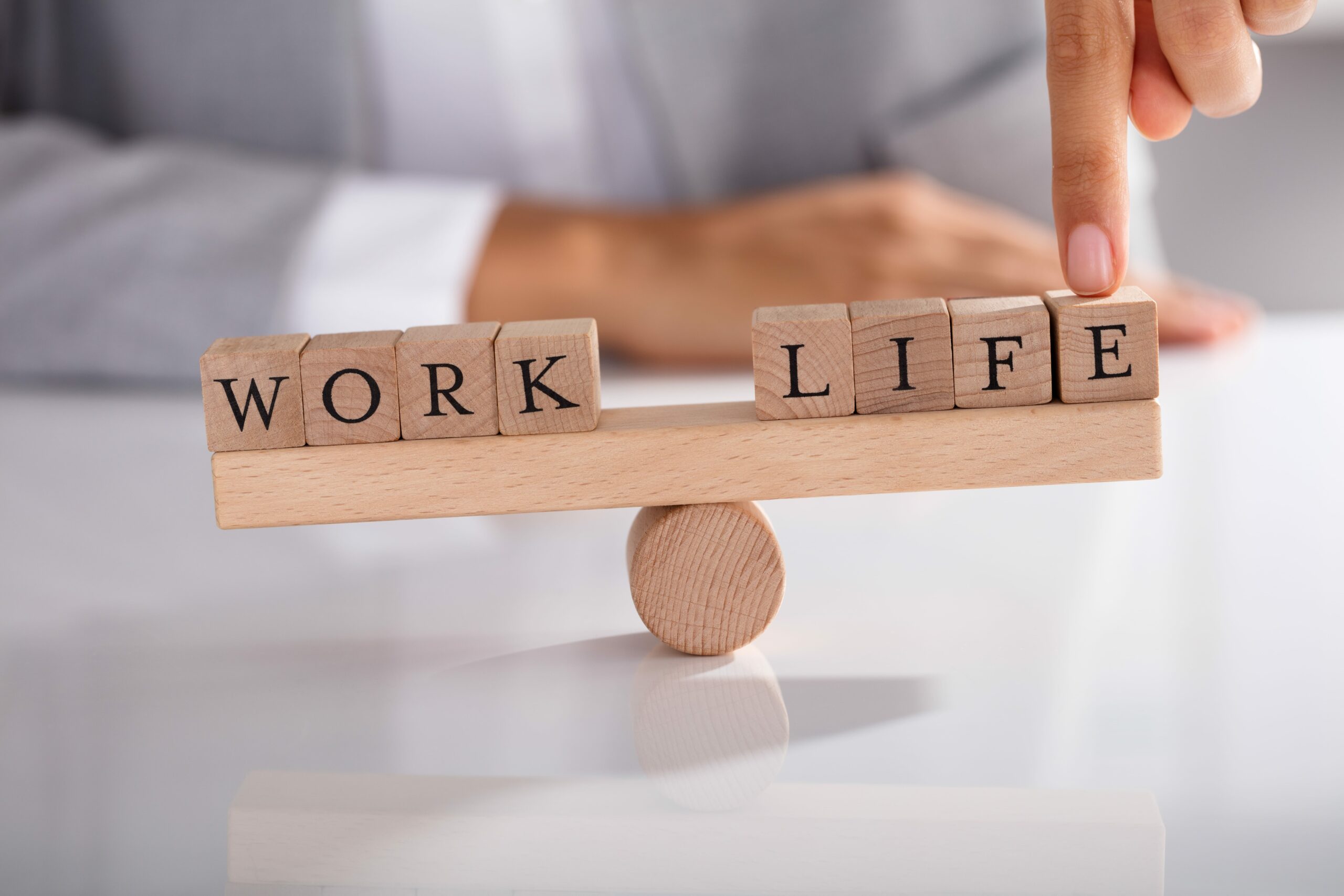 A person balances wooden blocks spelling WORK and LIFE on a seesaw, symbolizing juggling work and life. The hand adjusts the LIFE blocks, highlighting strategies for work-life balance. The blurred background emphasizes this delicate act.