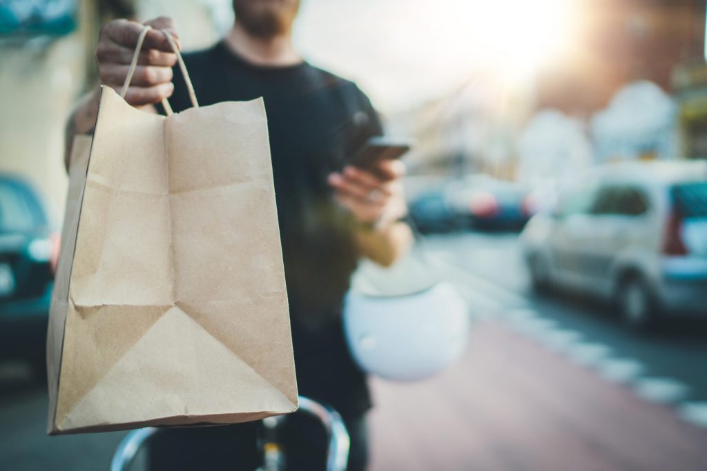 A person holding a brown paper bag in one hand while checking a smartphone with the other, embodying How to Thrive in the Gig Economy, stands outdoors on a city street with blurred cars and sunlight in the background.