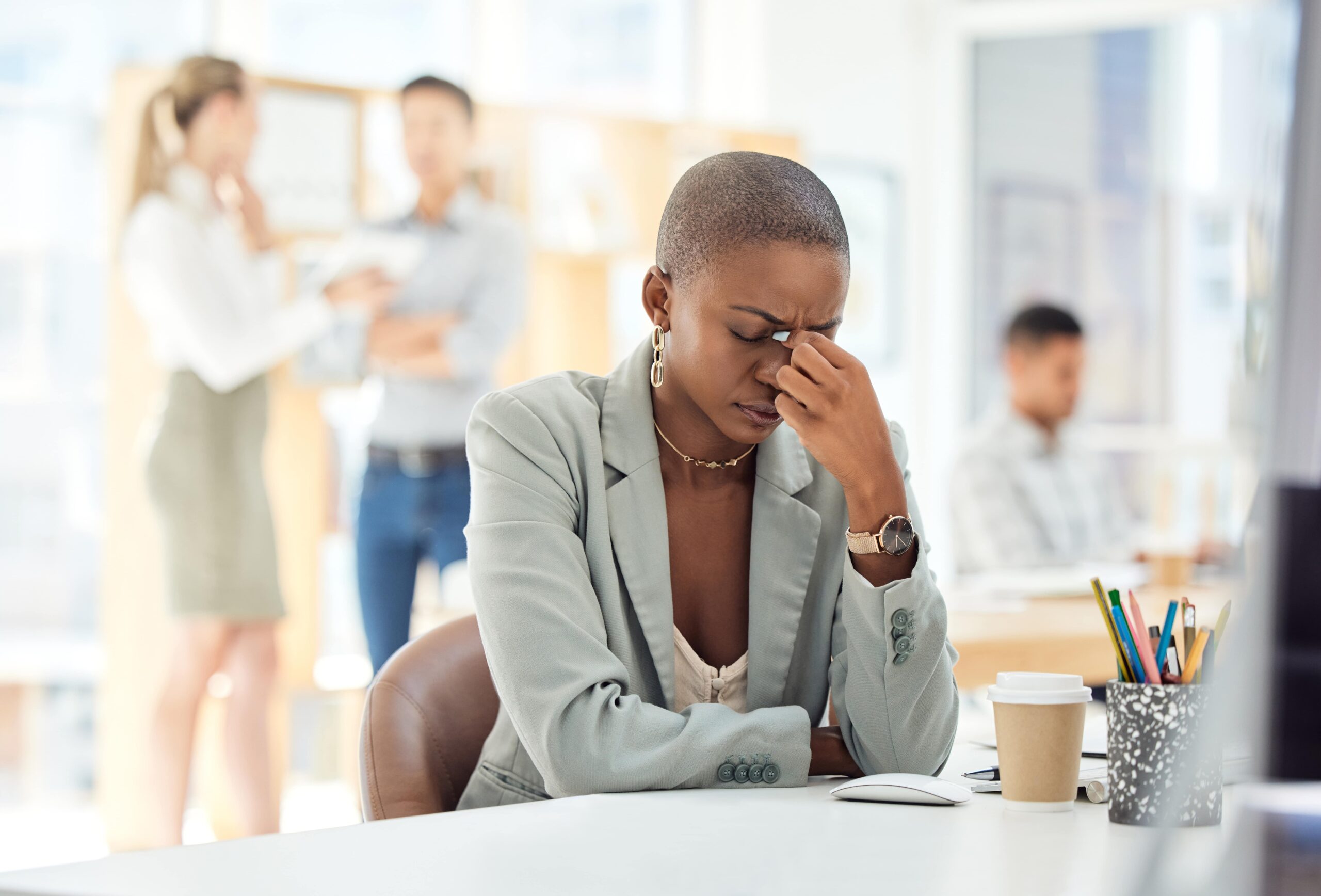 A woman in business attire sits at a desk, pinching the bridge of her nose in frustration—perhaps struggling to STOP Trying to Have it All and instead find a work-life balance that works for her as coworkers interact in the background.