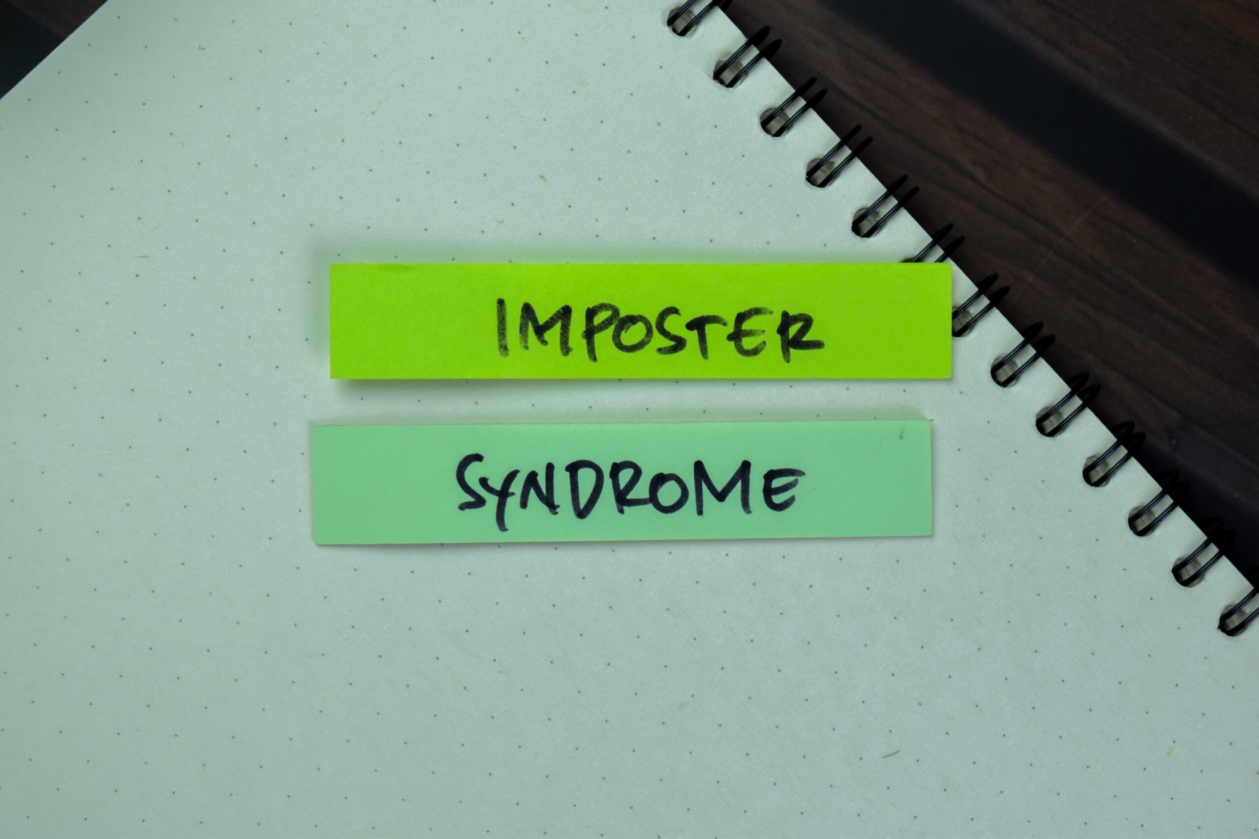 Two sticky notes on an open spiral notebook read IMPOSTER (yellow note) and SYNDROME (green note), with both words handwritten in black marker—perfect for illustrating tips on how to overcome imposter syndrome.