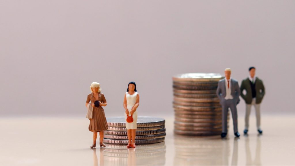 Miniature figures of two women stand before shorter stacks of coins, while two men stand behind taller stacks, highlighting gender disparity in earnings—an issue explored in Breaking the Glass Ceiling: How to Overcome Gender Stereotypes in Business.