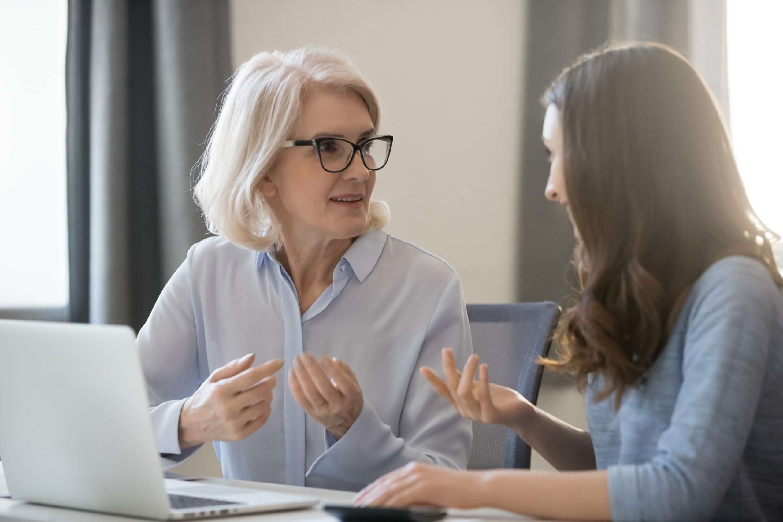 An older woman with gray hair and glasses talks to a younger woman at a desk with a laptop, both gesturing as they discuss the importance of finding a financial mentor and why every young woman should have one.