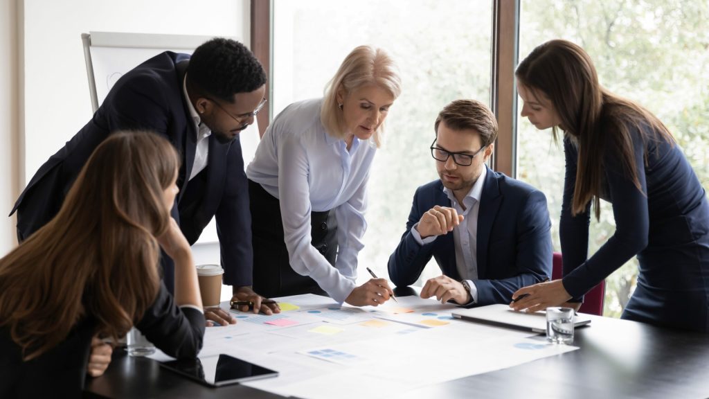 Five business professionals gather around a table, discussing documents and charts. One woman points at a paper while the others listen attentively—demonstrating Lead Like a Boss: Tips for Becoming a More Effective Leader in action. Natural light streams in behind them.