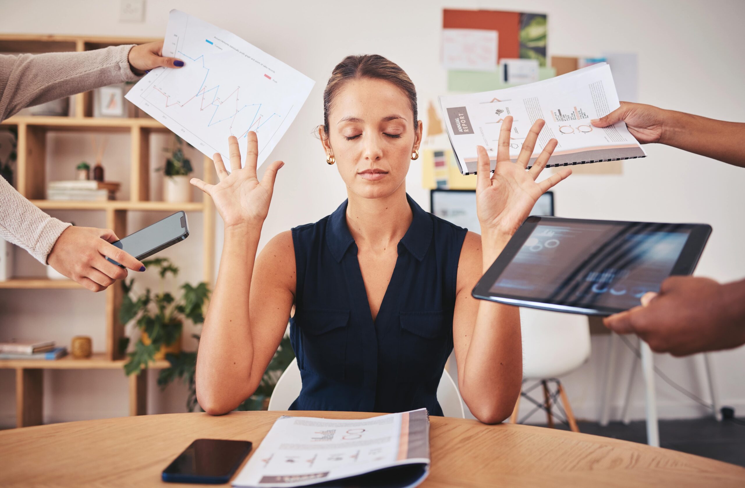 A woman sits calmly at her desk with eyes closed and hands raised, surrounded by phones and documents—showing how to manage your time effectively even in a hectic work environment.