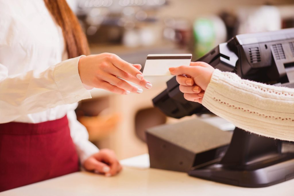 A cashier reaches to take a payment card from a customer at a checkout counter. Both hands are visible, with a register and payment terminal in the background.