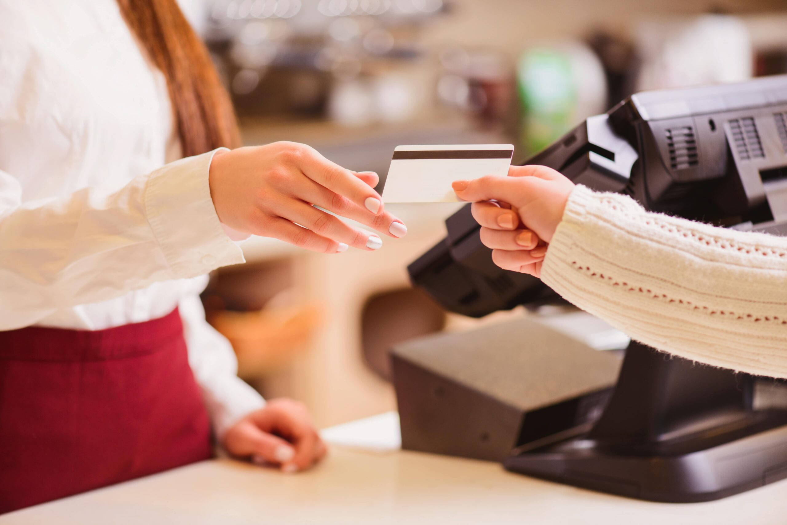 A cashier reaches to take a payment card from a customer at a checkout counter. Both hands are visible, with a register and payment terminal in the background.