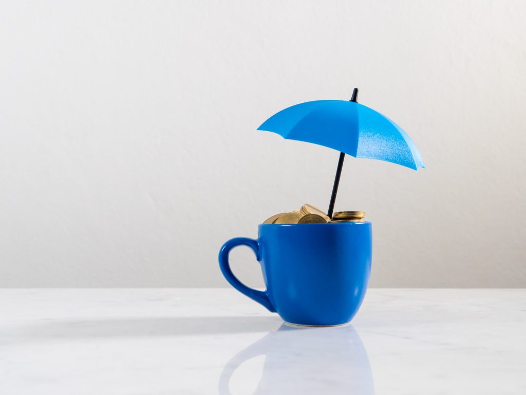 A small blue mug filled with coins sits on a white surface. A blue cocktail umbrella shelters them, symbolizing financial protection—an ideal image for “Emergency Funds and Rainy Days: Why Every Young Person Needs a Financial Safety Net.”.