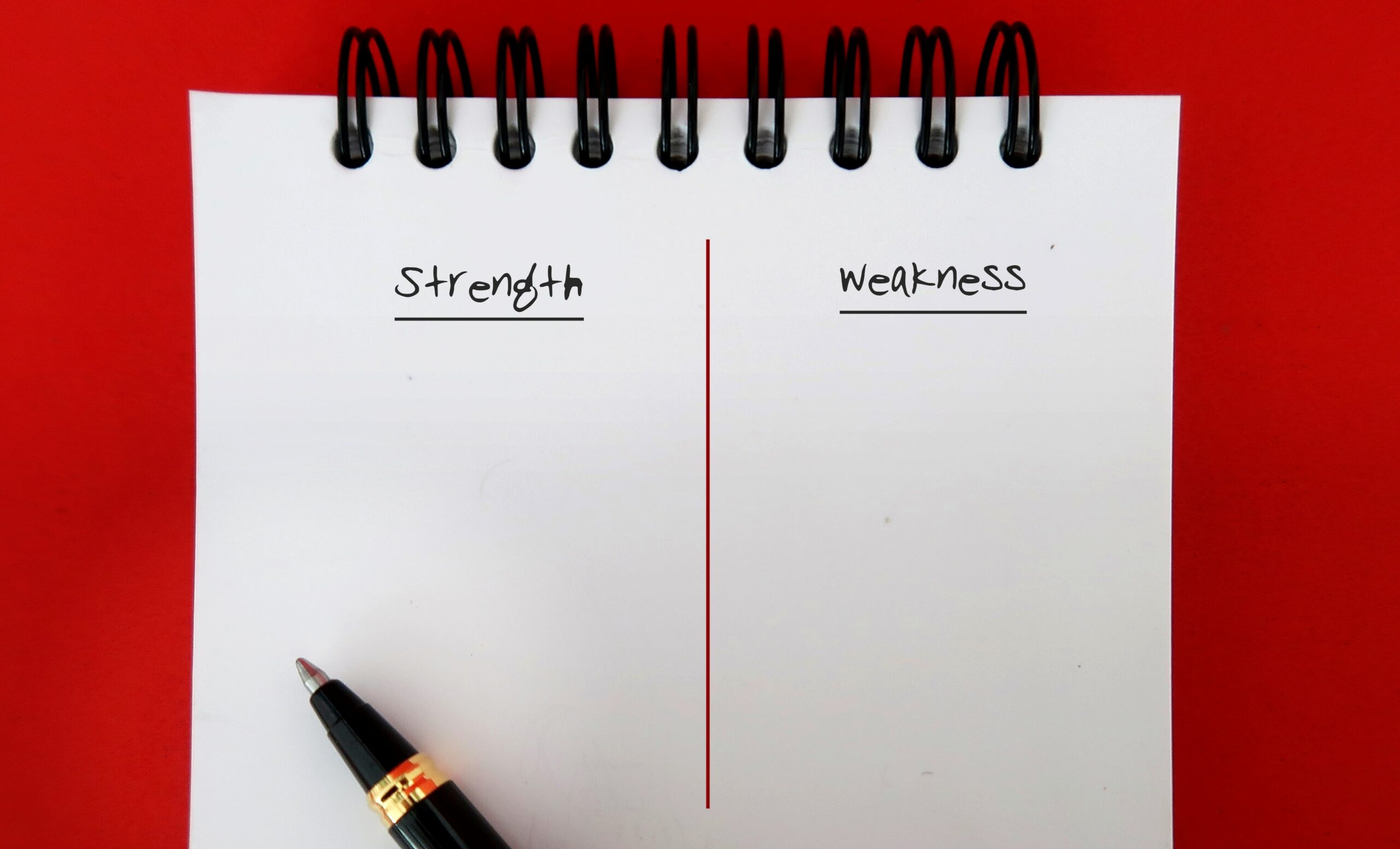 A notepad for self-assessment with Strength and Weakness columns, separated by a red line, rests on a red surface. Ideal for career development, it features a black pen positioned at the bottom left for identifying strengths and areas for growth.
