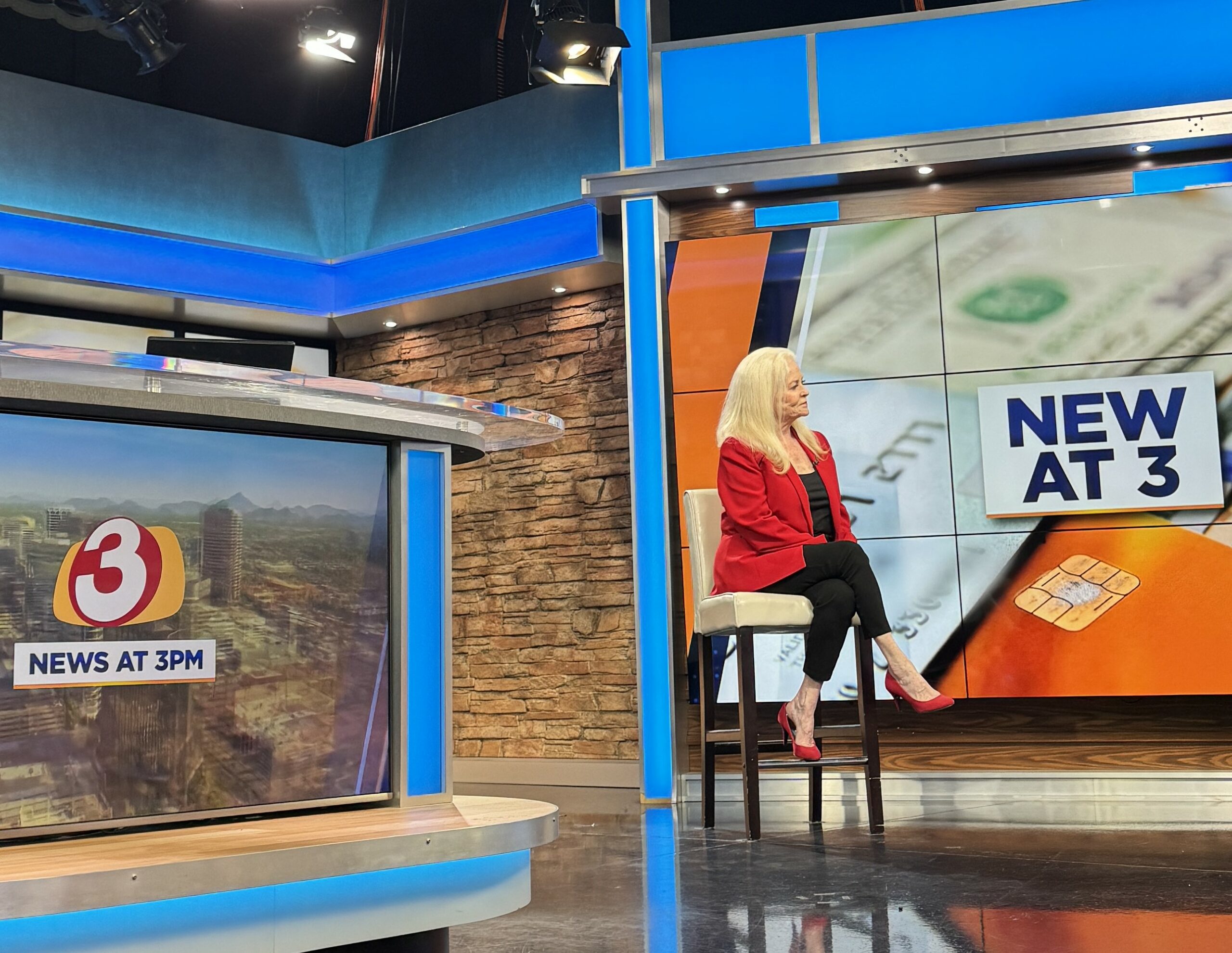 Sharon Lechter sits on a chair in a TV news studio, with “News at 3” displayed on screens behind her and on a news desk to the left. The set features stone and blue paneling.