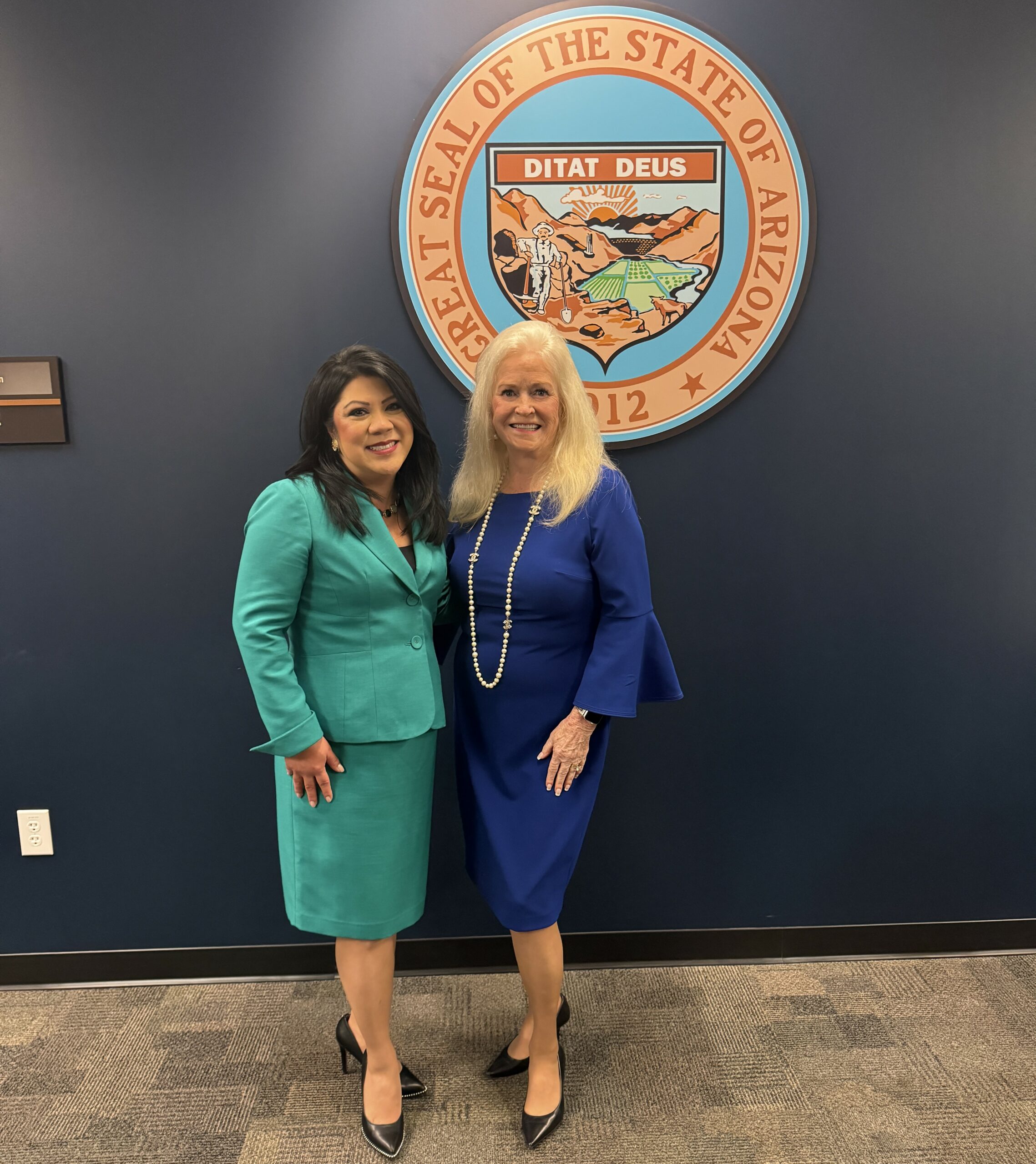 Two women stand smiling together in front of the Arizona state seal. One wears a teal skirt suit, and the other wears a royal blue dress with pearl jewelry. They are indoors on a carpeted floor.