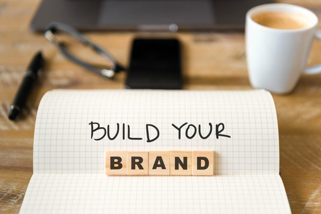 A notebook with the words BUILD YOUR written on grid paper and BRAND spelled out with wooden letter tiles, illustrating The Power of Personal Branding. A pen, glasses, smartphone, and coffee cup rest on the wooden desk in the background.