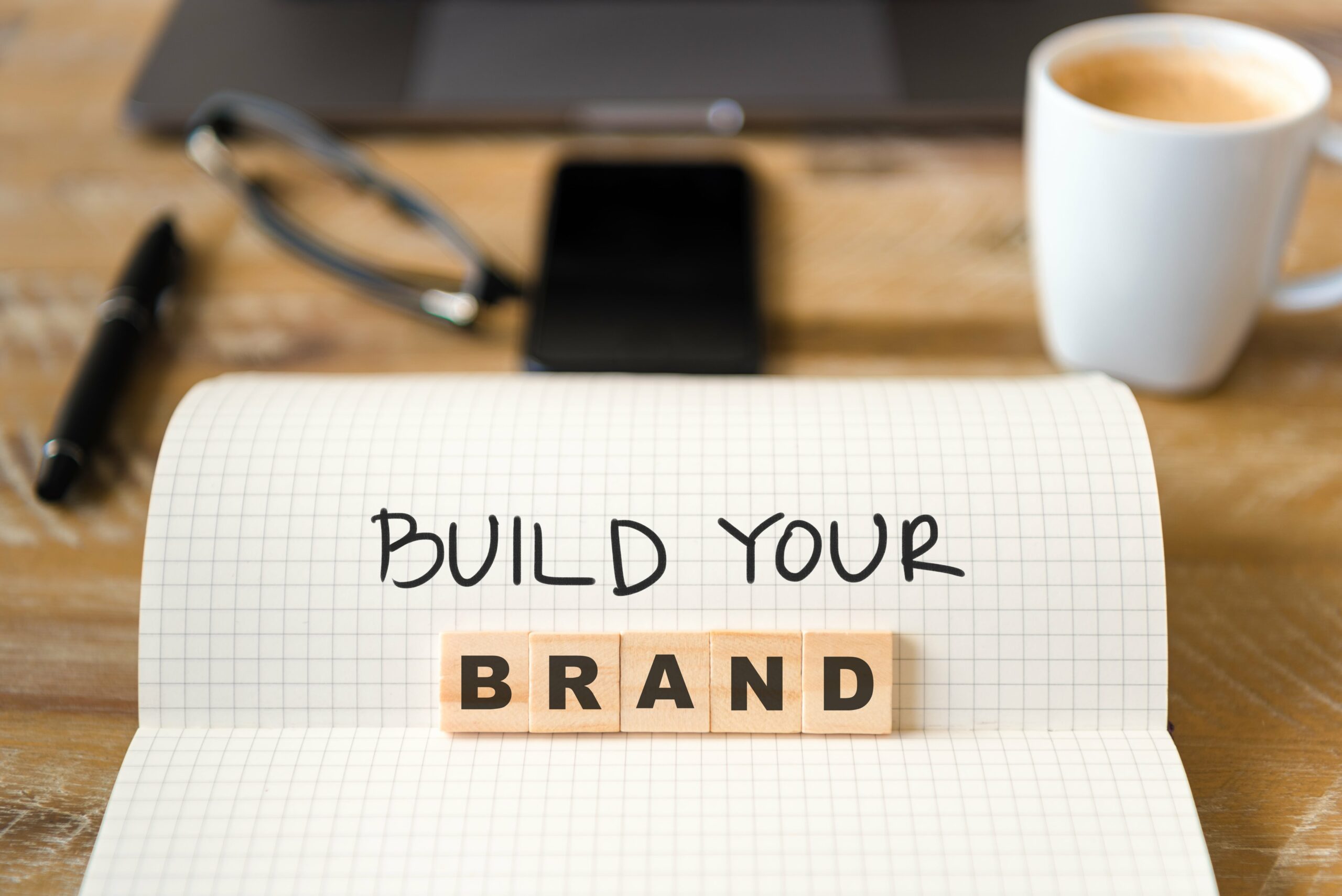 A notebook with the words BUILD YOUR written on grid paper and BRAND spelled out with wooden letter tiles, illustrating The Power of Personal Branding. A pen, glasses, smartphone, and coffee cup rest on the wooden desk in the background.