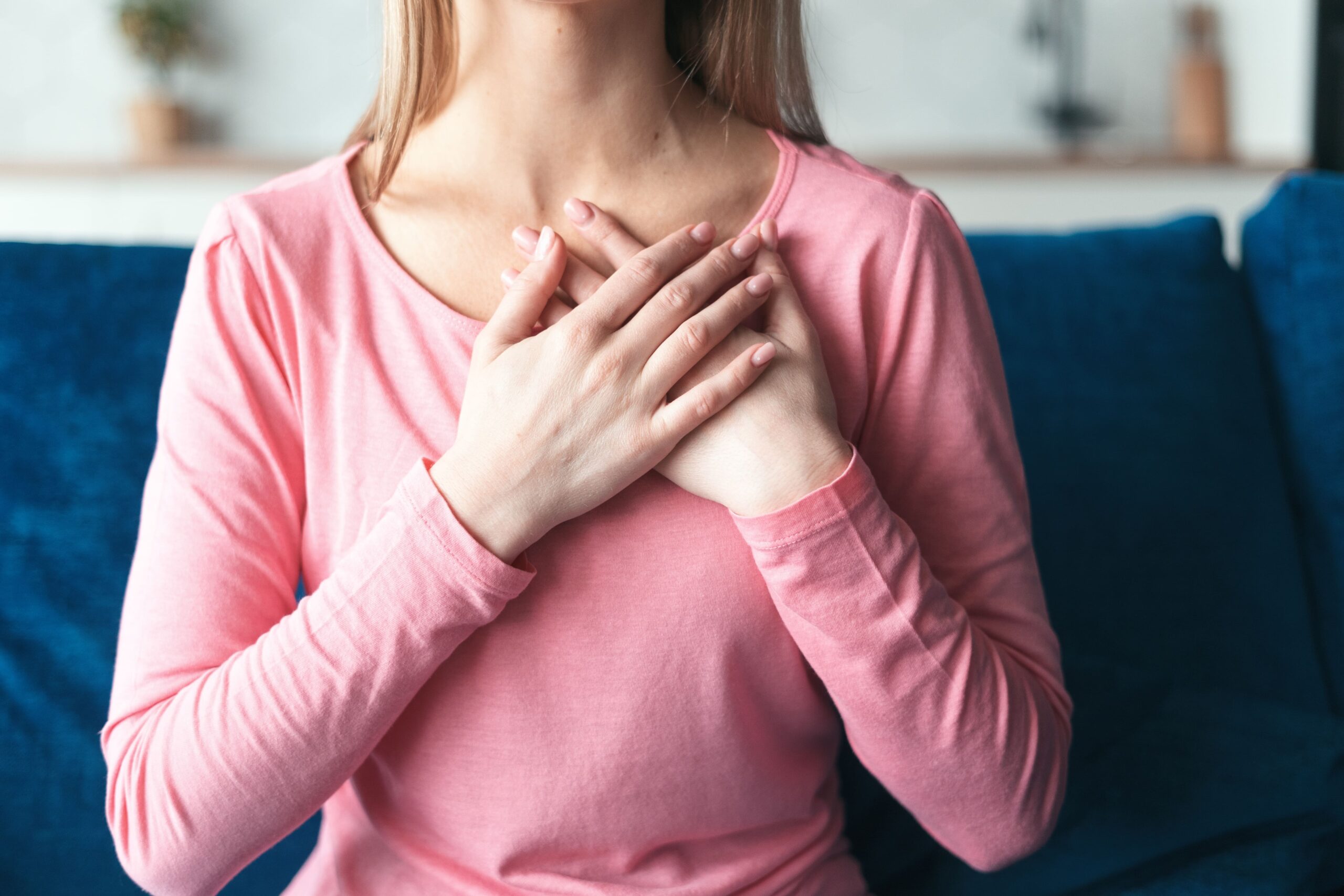 A woman in a pink long-sleeve shirt sits on a blue sofa with both hands over her chest, embodying self-compassion—an essential step in getting past failure to create success.