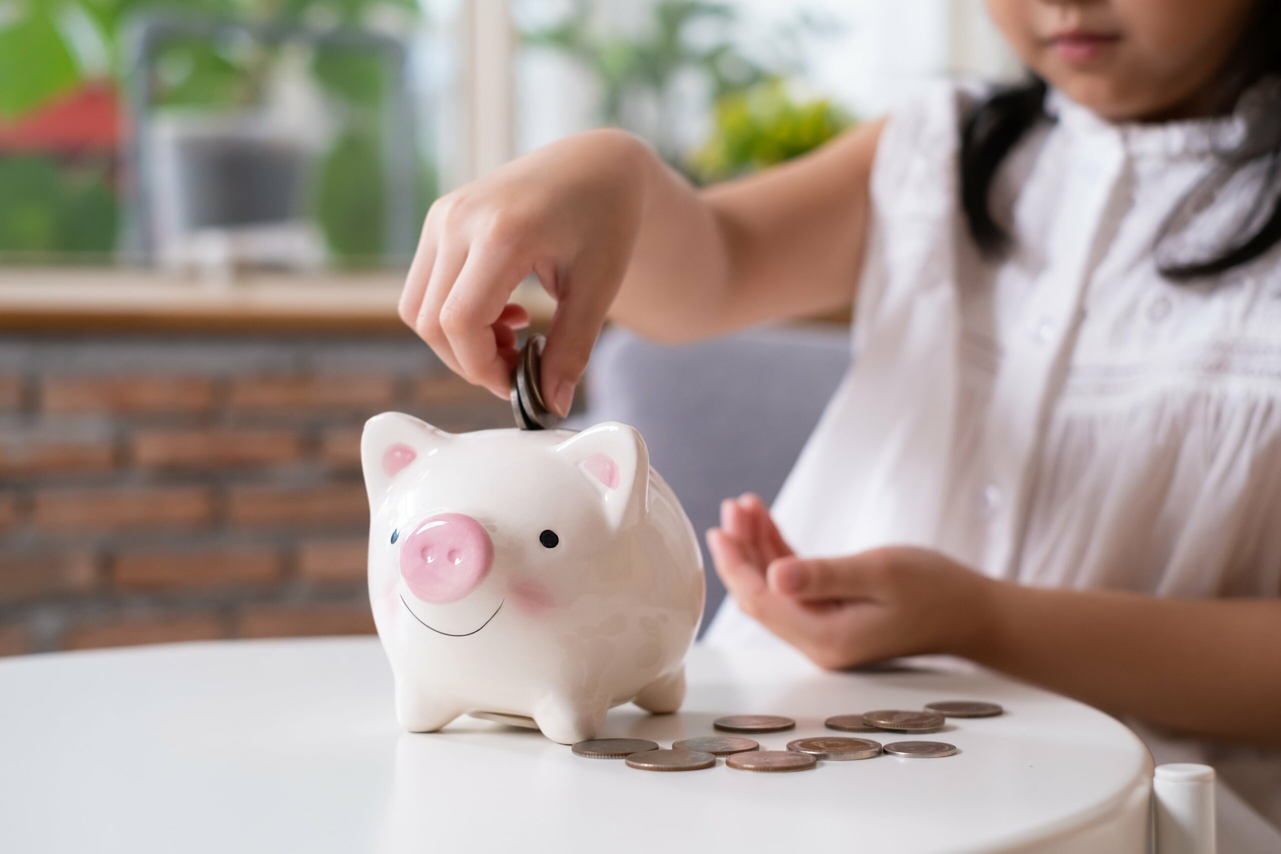 A young girl in a white shirt puts a coin into a white piggy bank on a table, with several coins scattered beside it—Raising Smart Savers starts early, making every moment a guide to teaching your child financial literacy.