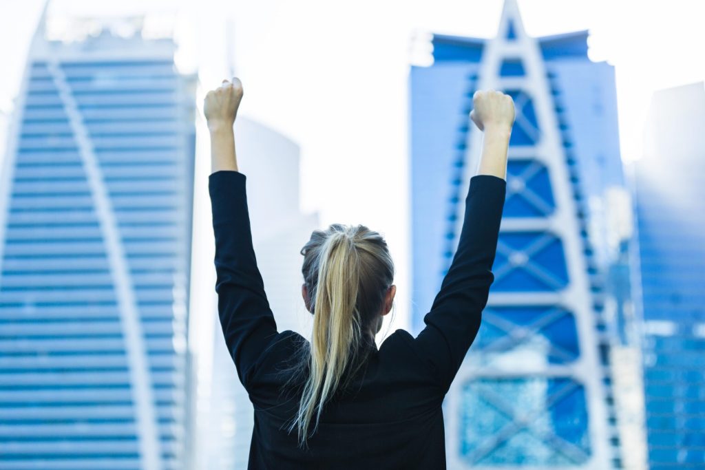 A woman in a black blazer stands with her back to the camera, raising both arms in celebration against city skyscrapers—an image of Achieving Personal and Professional Success.