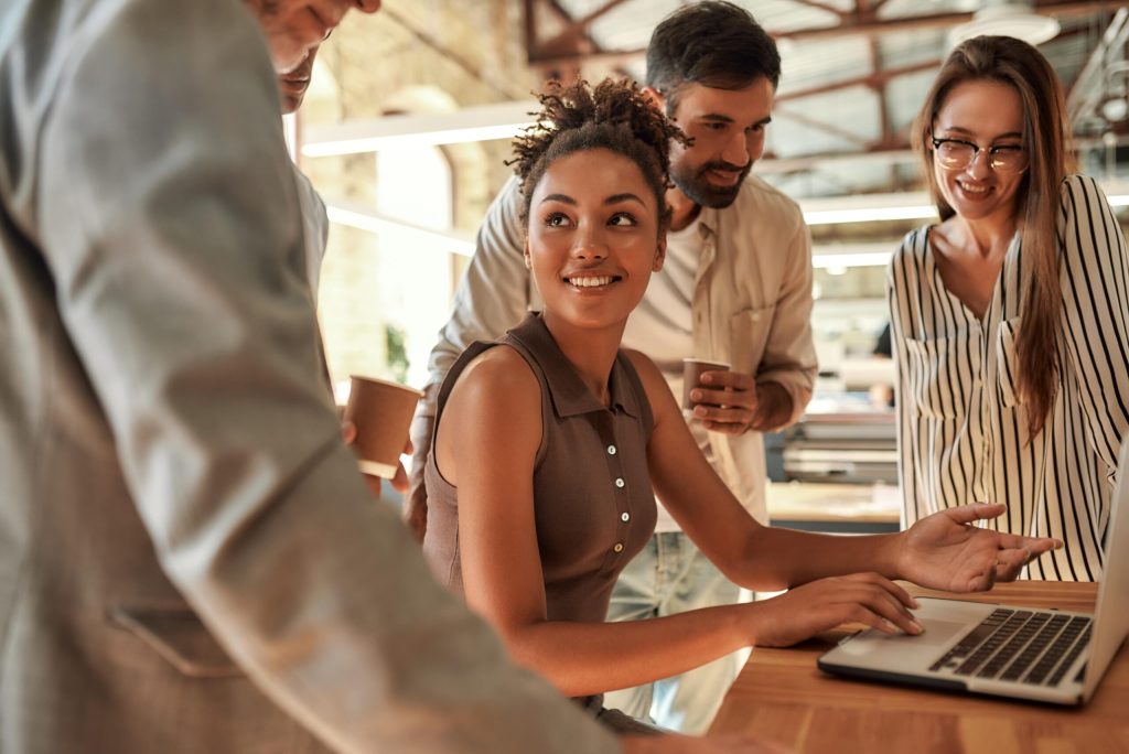 A group of four people gathers around a desk, smiling and talking as one woman sits at a laptop. The modern, casual office with warm lighting reflects how young professionals boost energy and productivity by thriving in the fast lane.
