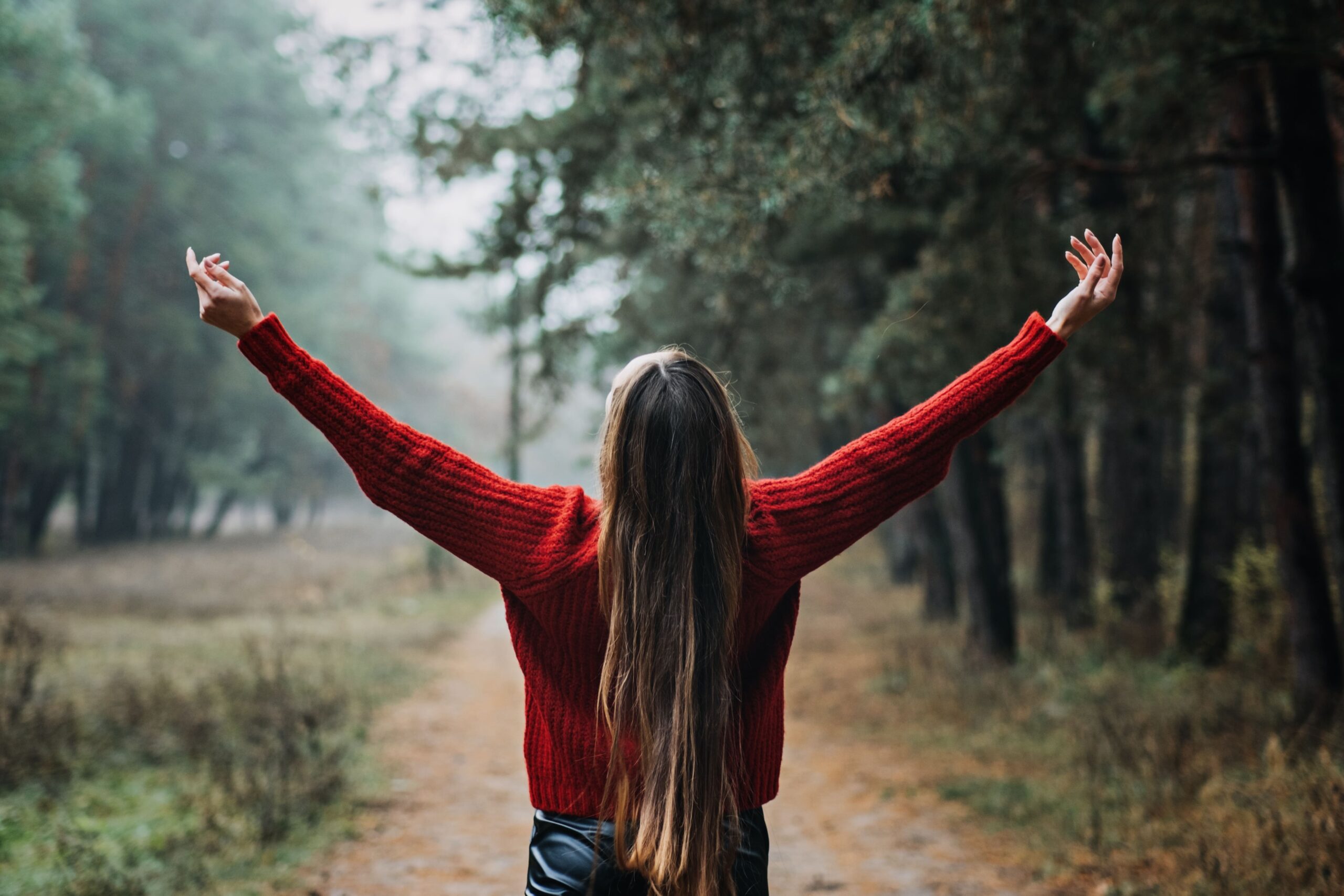 A woman with long hair in a red sweater stands on a forest path, arms raised, embracing the misty woodsโembodying Forging Resilience: The Secrets for Developing an Indestructible Inner Strength.