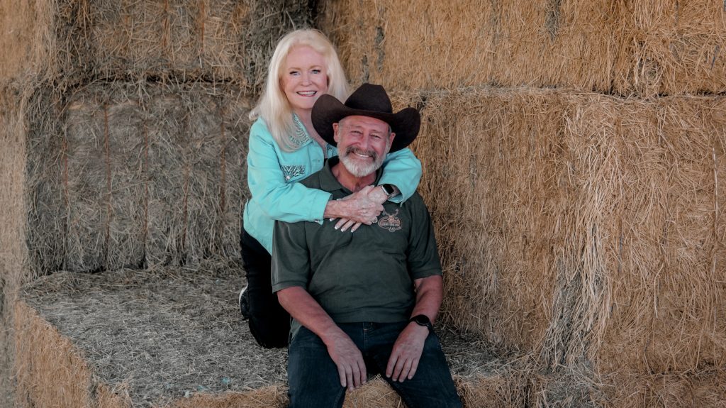 A smiling woman with long blonde hair embraces a bearded man wearing a cowboy hat. They are sitting together on stacked hay bales in a barn setting.
