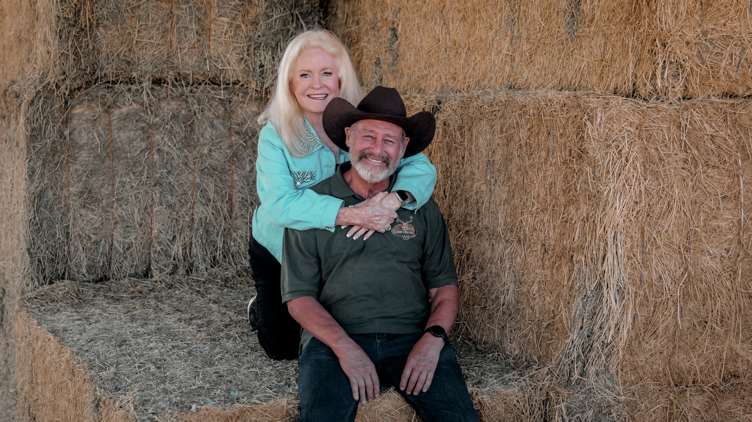 A smiling woman with long blonde hair embraces a bearded man wearing a cowboy hat. They are sitting together on stacked hay bales in a barn setting.
