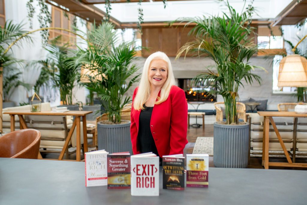 A smiling woman with long blonde hair, wearing a bright red blazer, stands behind a table displaying five business books in a spacious room filled with plants and modern decor, ready to share Money Saving Tips on Amazon Prime Day.