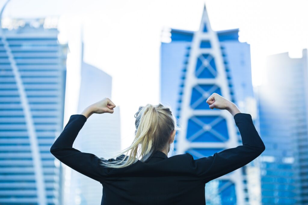 Woman in a suit flexing her arm muscle in front of blue skyscrapers.