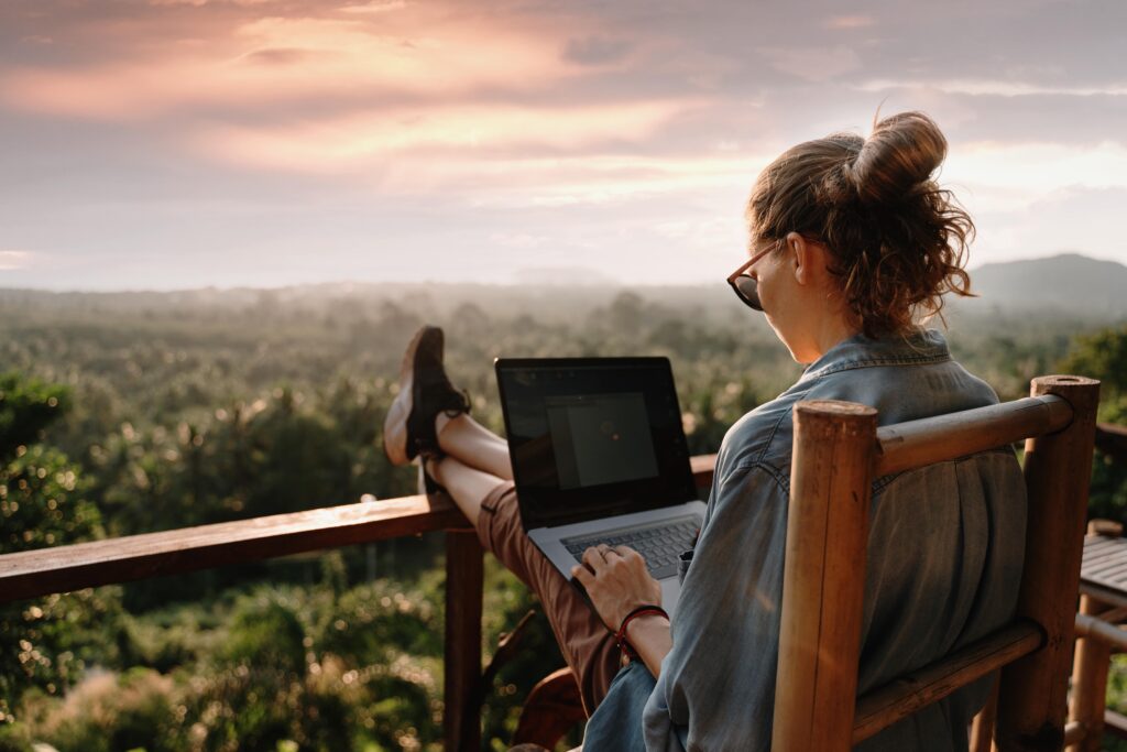 A freelancer sitting on a deck working on her laptop looking over a forest.