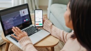 A woman working on her laptop and mobile device, looking at cryptocurrency and stock trends. 