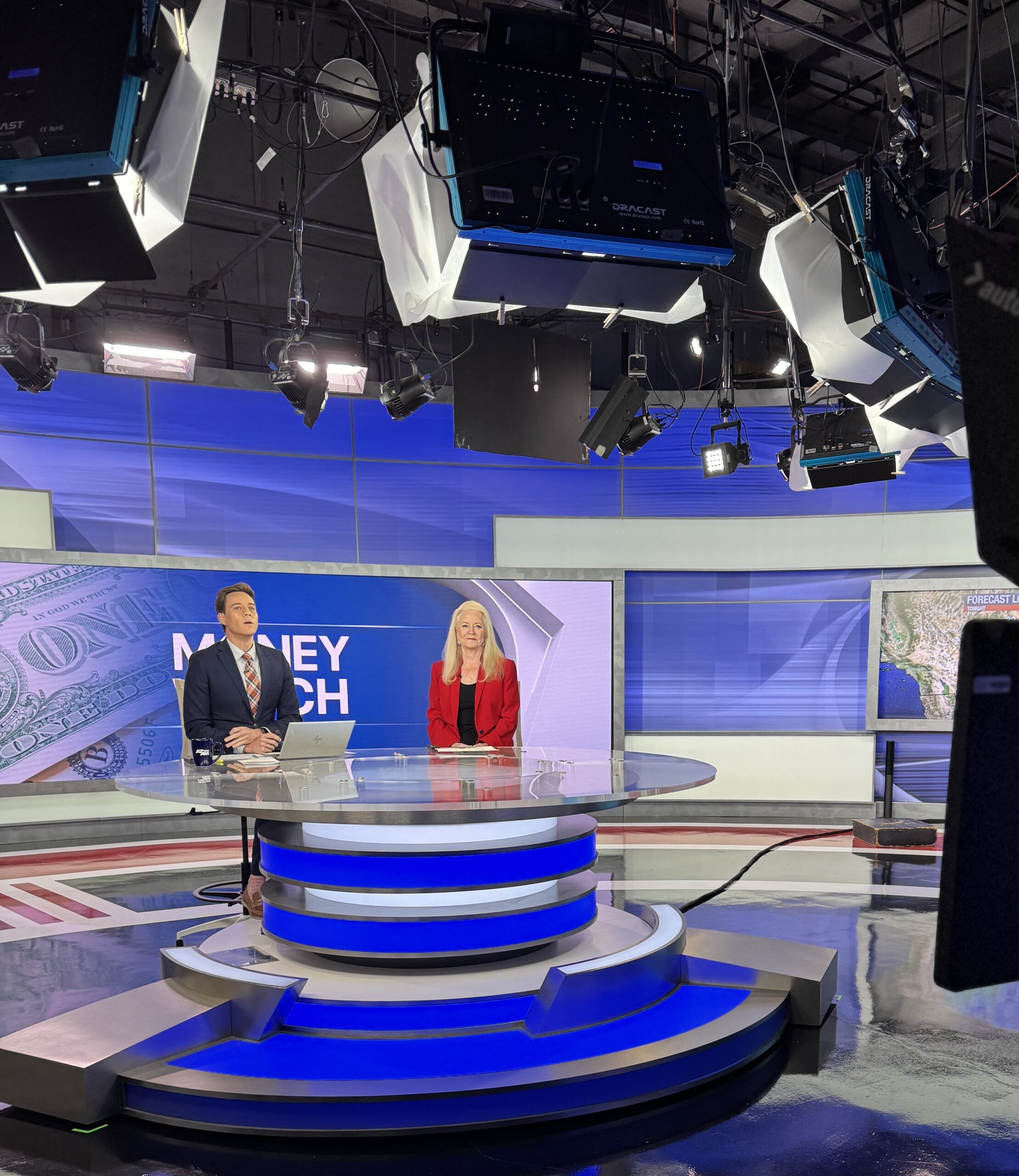 Sharon Lechter sits at a round glass desk with a news anchor in a brightly lit TV studio. Large monitors, lights, and a screen displaying “Money Watch” and Money Saving Tips on Amazon Prime Day are visible in the background.