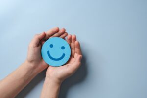 An individual's hands reaching out and holding a blue cutout smiley face over a blue background.