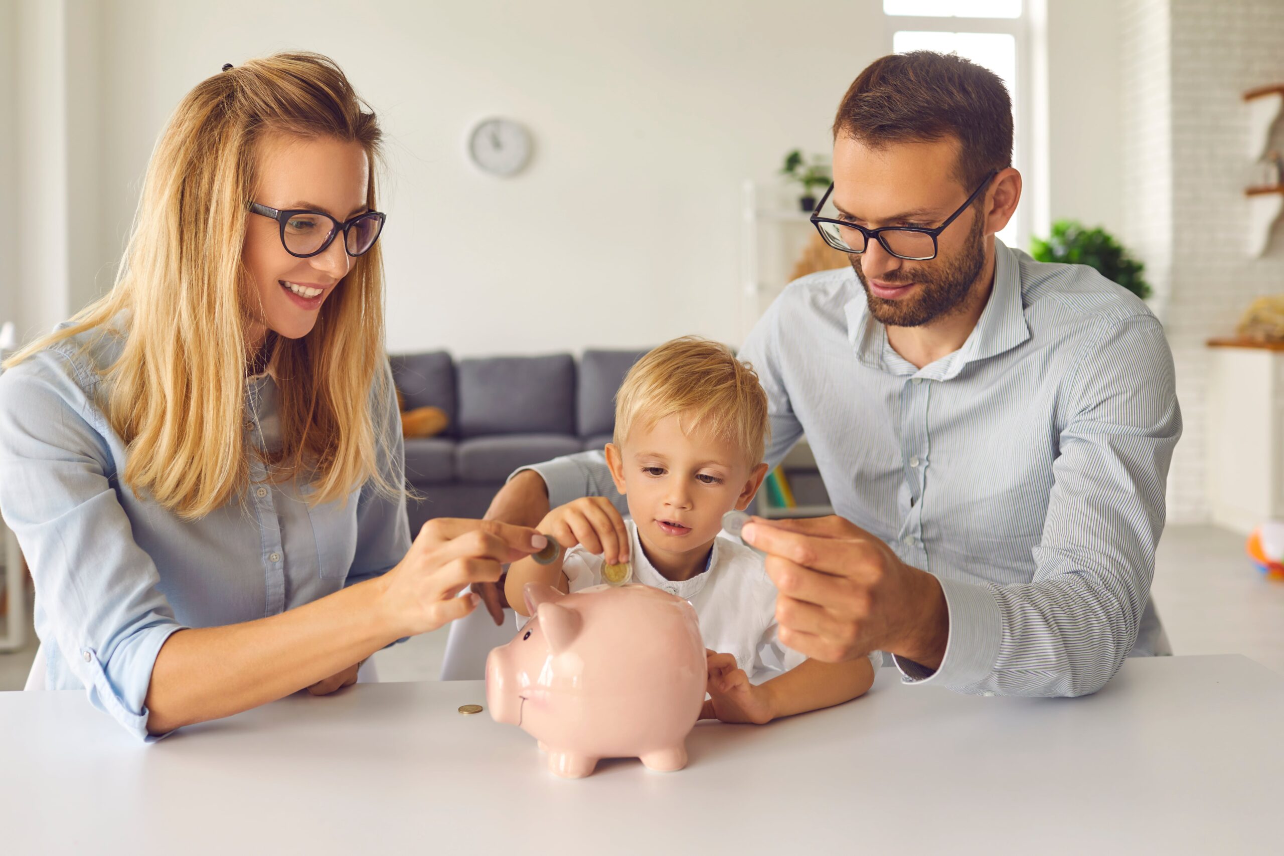 Parents teaching their young son how to save money by putting coins in a piggy bank at home. 