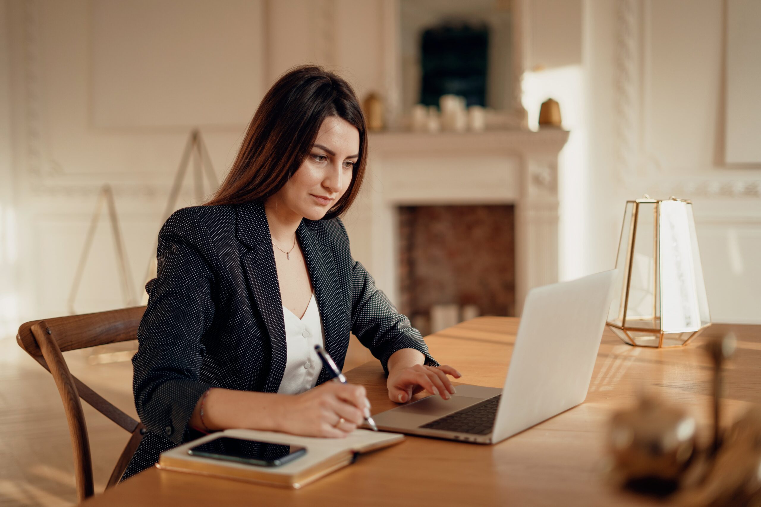 Professional young woman in a blazer working on a laptop while taking notes at a wooden desk in a ​​well-lit, elegantly decorated room.
