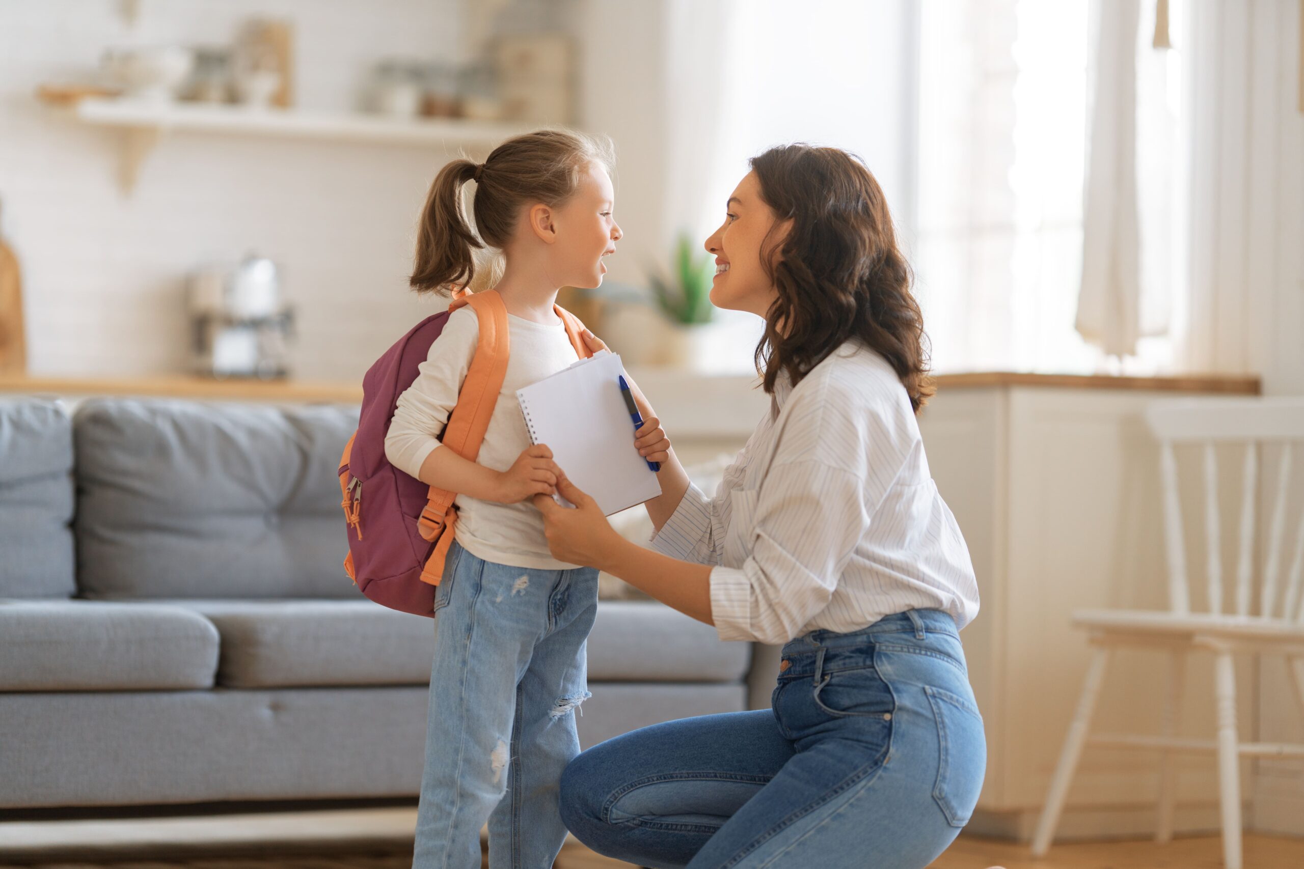 Mother smiling and encouraging her daughter who is wearing a backpack and holding a notebook, preparing for school.