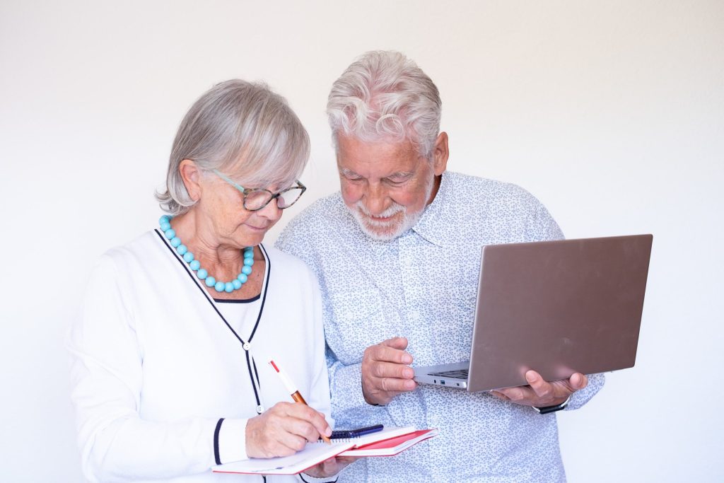 Senior couple working on their finances—woman writing in a notebook while the man holds a laptop, both engaged and smiling.