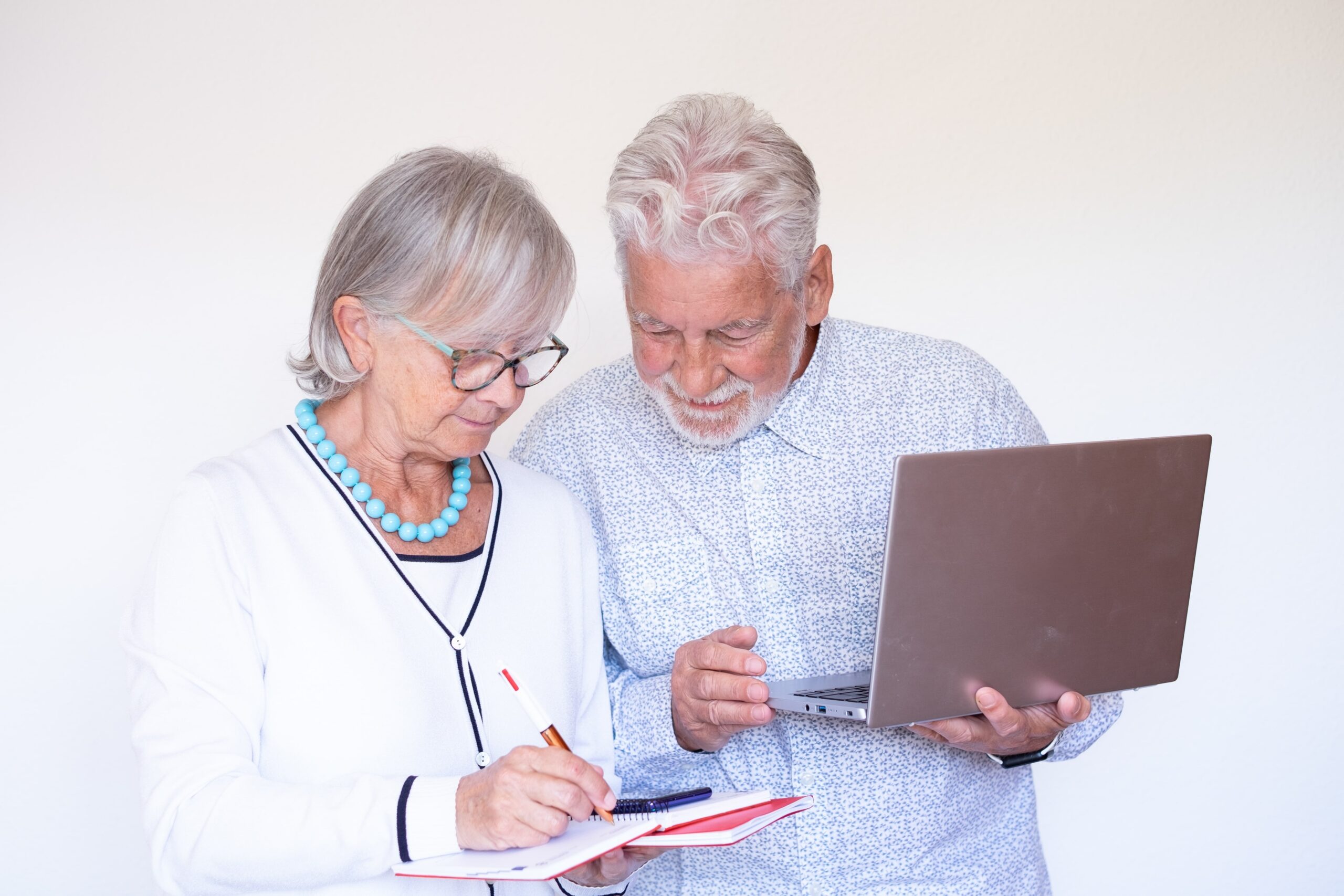 Senior couple working on their finances—woman writing in a notebook while the man holds a laptop, both engaged and smiling.