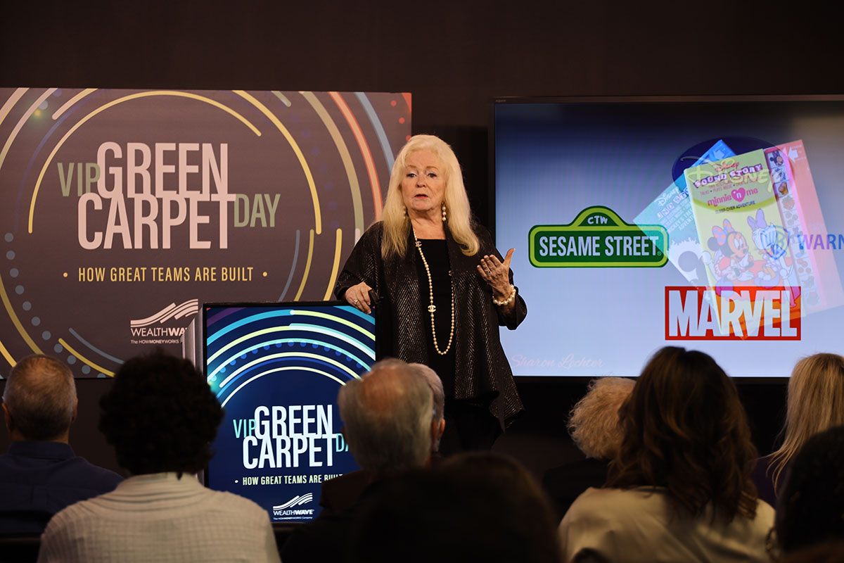 A woman speaks on stage to an audience, standing between two screens—one reads “VIP Green Carpet Day” and the other displays logos for Sesame Street, Marvel, and other brands.