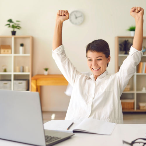 Smiling woman sitting at a desk with a laptop and notebook, raising her arms in celebration while working in a bright home office