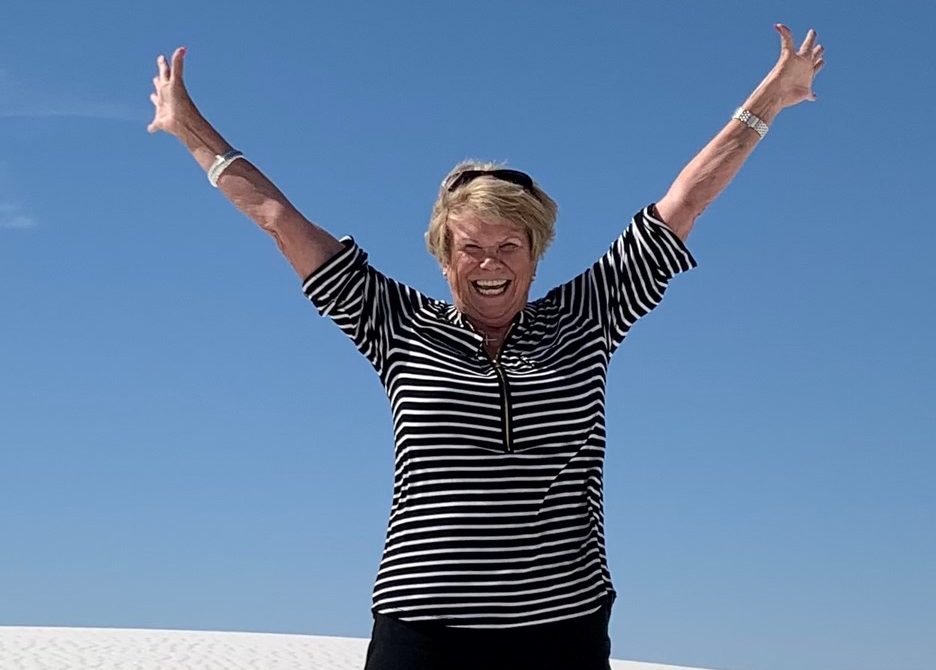Smiling woman standing on white sand with arms raised high, wearing a black and white striped shirt and black pants, against a clear blue sky.
