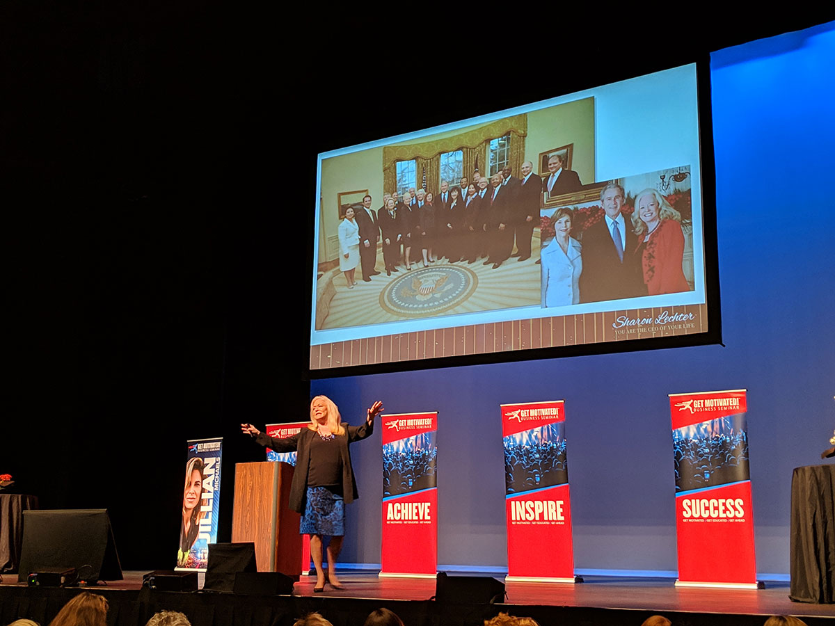 Sharon Lechter on stage in front of a large screen displaying an Oval Office photo. Banners reading "ACHIEVE," "INSPIRE," and "SUCCESS"