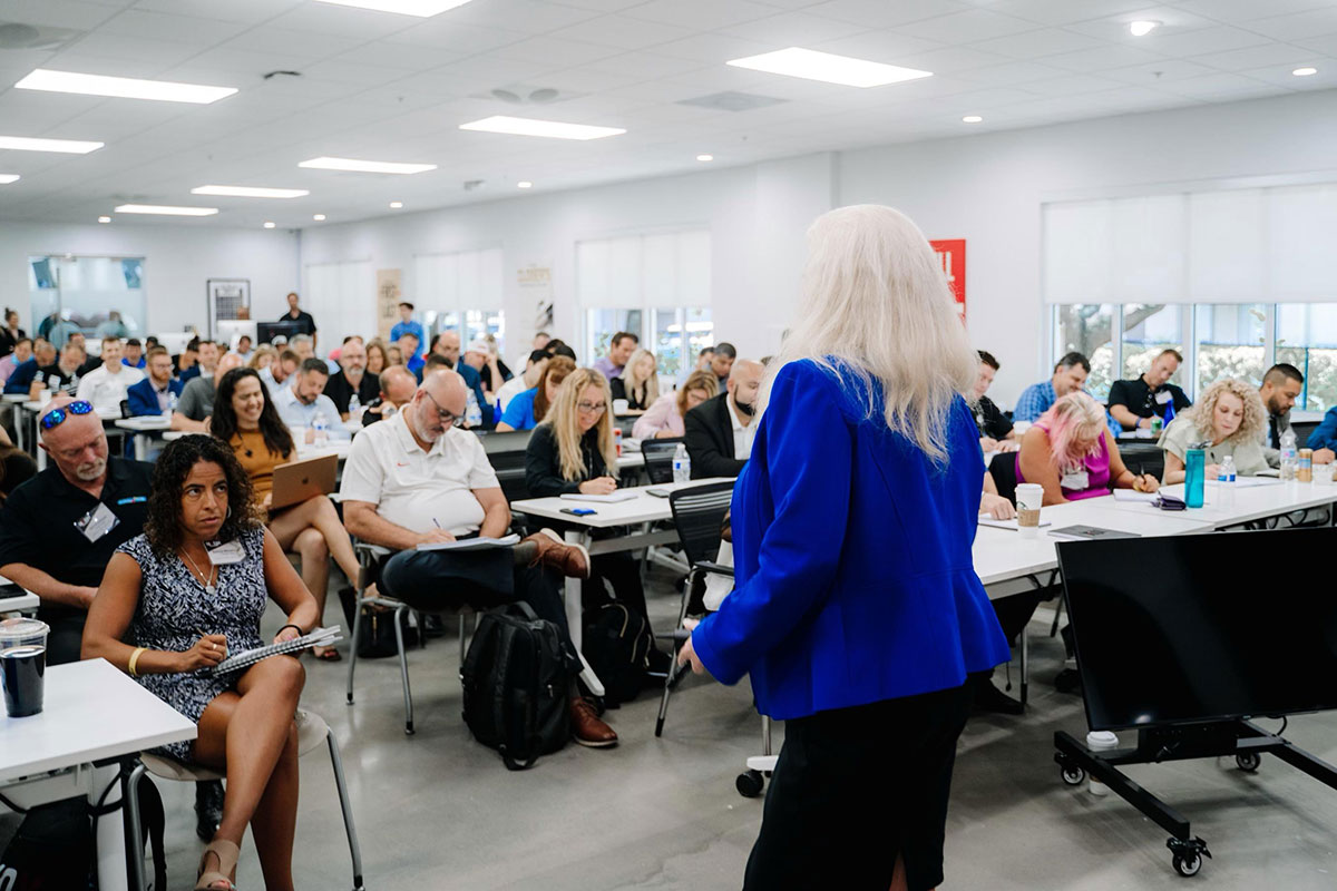 Sharon Lechter in a blue blazer addresses a classroom of diverse adults seated at tables, taking notes