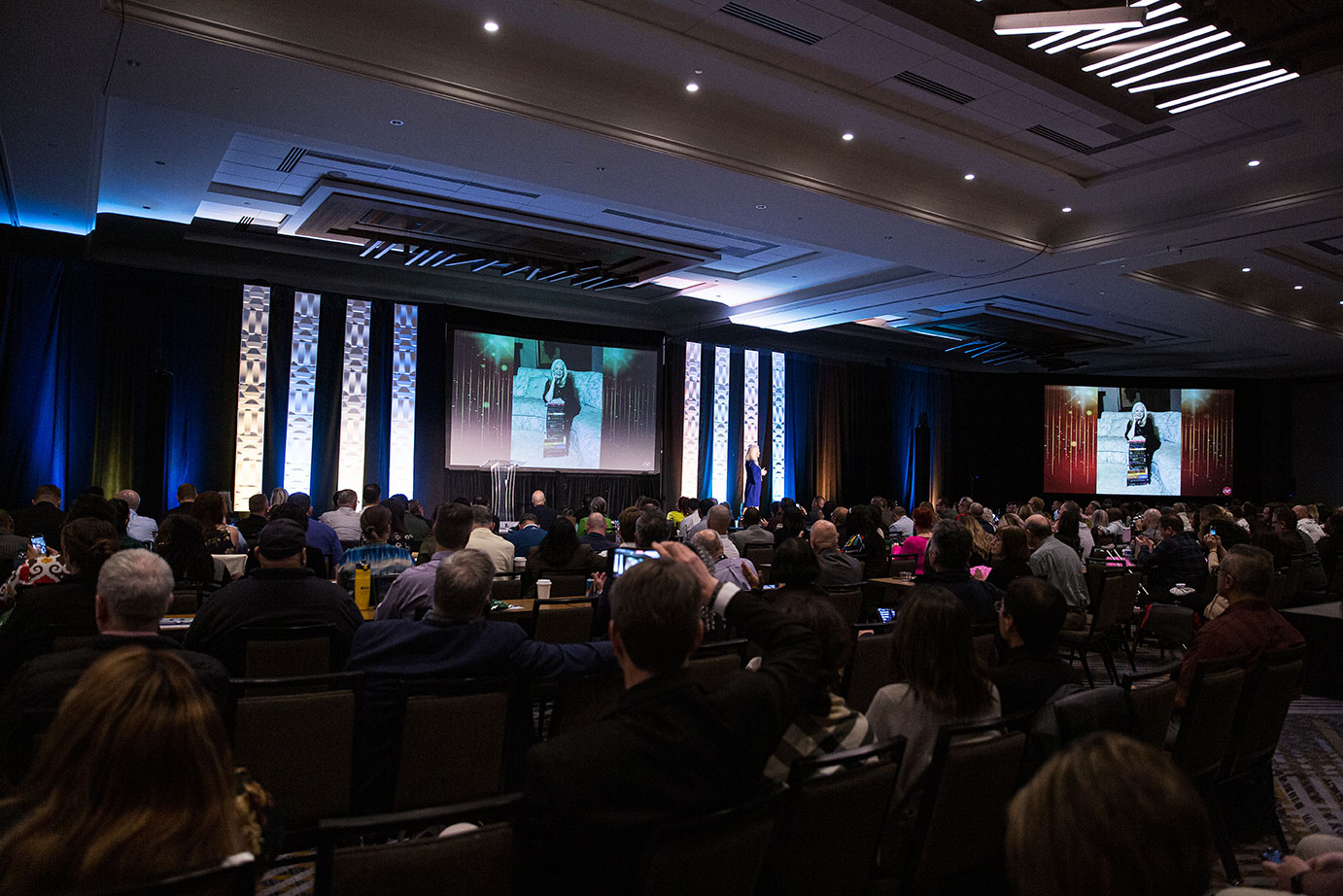 Audience seated in a large conference room, with Sharon Lechter speaking