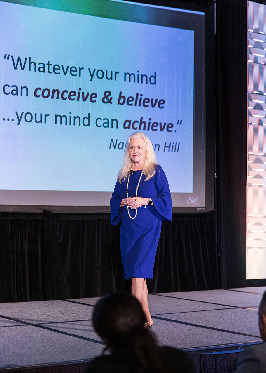 Sharon Lechter in blue speaks confidently on stage. Behind her, a screen displays the quote: Whatever your mind can conceive & believe... your mind can achieve